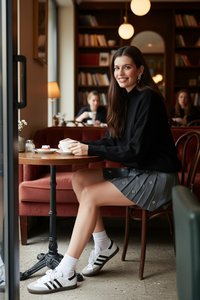Mujer con suéter negro y falda gris sentada en una mesa redonda en una cafetería, sosteniendo una taza blanca, sonriendo, con estanterías de libros al fondo.