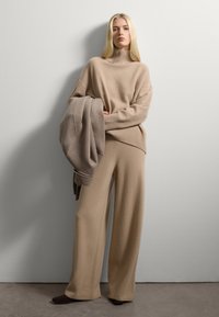 Pull en tricot beige avec un col montant, associé à un pantalon large. Texture douce, coupe oversize et une écharpe assortie drapée sur un bras.