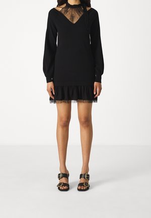 Vrouw draagt een zwarte jurk met lange mouwen, doorschijnende kanten halslijn en zoom, gecombineerd met zwarte open-toe sandalen met gouden gespen.