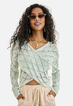 Mujer con cabello rizado que lleva gafas de sol estilo cat-eye, collar de oro, suéter de punto verde claro con patrón de diamantes y pantalones beige con cordón ajustable.