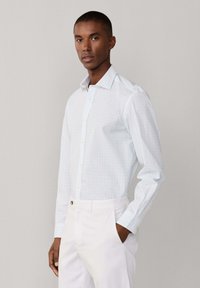 Homme portant une chemise à carreaux bleu clair et un pantalon blanc, debout avec une main dans la poche devant un fond uni.