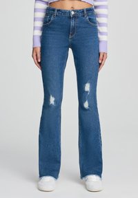 Jeans svasati blu in denim con vita alta, caratterizzati da toppe strappate e orli sfilacciati, abbinati a sneaker bianche.