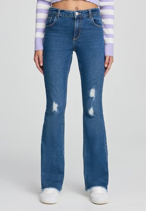 Jeans svasati blu in denim con vita alta, caratterizzati da toppe strappate e orli sfilacciati, abbinati a sneaker bianche.