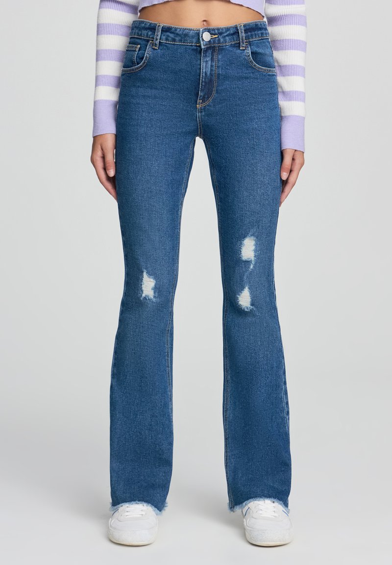 Jeans svasati blu in denim con vita alta, caratterizzati da toppe strappate e orli sfilacciati, abbinati a sneaker bianche.