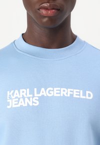 Šviesiai mėlynas džemperis, pagamintas iš minkštos medžiagos, turi suapvalintą kaklą ir baltą "KARL LAGERFELD JEANS" logotipą ant krūtinės.