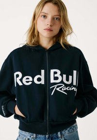Crna zip jakna od mekog materijala s natpisom "Red Bull Racing" ispisanim bijelim podebljanim slovima na prednjoj strani. Sadrži rebraste manžete i pojas.
