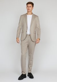 Costum bej deschis cu un finisaj texturat, sacou cu un singur rând de nasturi și pantaloni cu fața plată, asortat cu un tricou alb și pantofi negri.