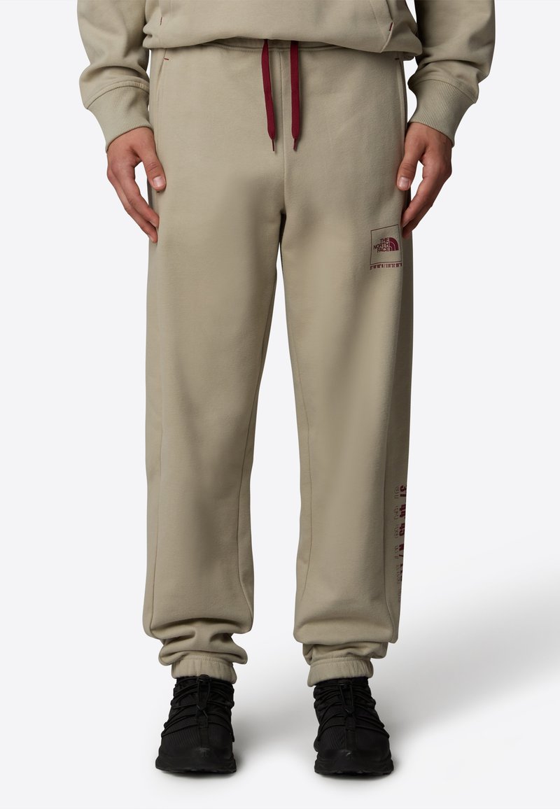 Pantalones de chándal beige hechos de una tela suave, con puños elásticos, un cordón rojo y estampados de logo en la parte delantera y lateral.