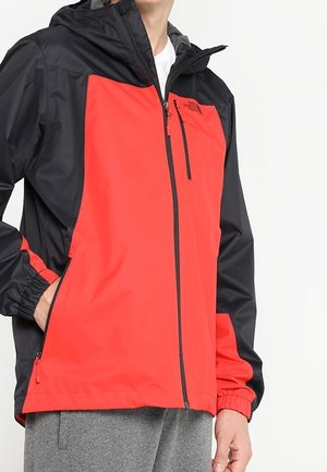 Person trägt eine schwarz-rote Outdoorjacke mit Reißverschluss, Kapuze und elastischen Bündchen, mit einer Hand in der Tasche.