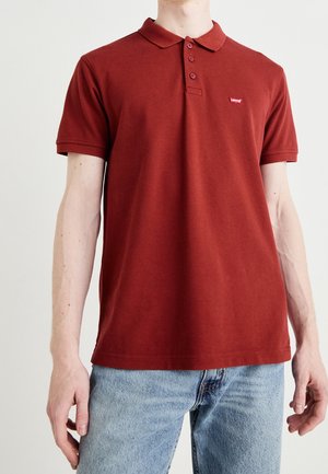Person trägt ein rotes Poloshirt mit kurzen Ärmeln, drei Knöpfen und kleinem Levi's-Logo auf der Brust, kombiniert mit blauen Jeans.