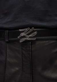 Zwarte leren riem met een zilveren metalen gesp in een vloeiend, cursief ontwerp. De riem heeft een gladde textuur en nette stiksels.