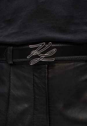 Zwarte leren riem met een zilveren metalen gesp in een vloeiend, cursief ontwerp. De riem heeft een gladde textuur en nette stiksels.
