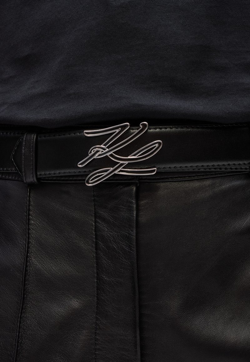 Zwarte leren riem met een zilveren metalen gesp in een vloeiend, cursief ontwerp. De riem heeft een gladde textuur en nette stiksels.