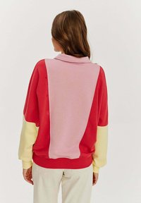 Sweatshirt s barevnými bloky, které obsahují červené, růžové a žluté sekce, s jemnou texturou a designem s polo límečkem.