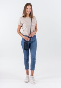 Jonge vrouw die staat, glimlacht, een beige t-shirt draagt, blauwe cropped jeans, witte sneakers en een kleine zwarte crossbodytas tegen een witte achtergrond.