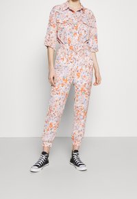 Mono floral en rosa suave con patrones en naranja, verde y azul. Presenta mangas cortas, cinturón en la cintura y puños elásticos en los tobillos.