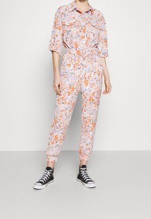 Bloemen jumpsuit in zachtroze met oranje, groene en blauwe patronen. Heeft korte mouwen, een riem op de taille en elastische manchetten bij de enkels.