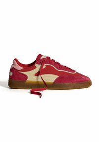 Red and beige suede sneakers with a gum rubber sole, featuring red laces, side cutouts, and a textured upper design.