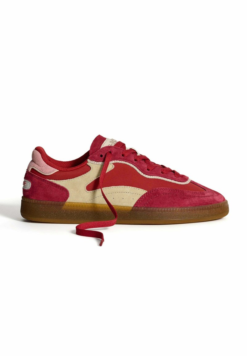 Red and beige suede sneakers with a gum rubber sole, featuring red laces, side cutouts, and a textured upper design.