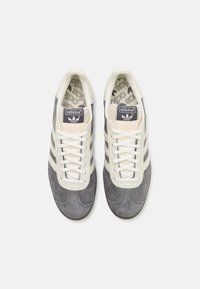 Tênis de camurça cinza com detalhes em creme, atacadores brancos e três riscas cinzas. A palmilha apresenta um padrão repetido de log�ótipos.