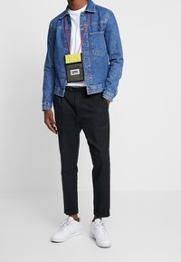 Giacca di jeans azzurro chiaro con tasche sul petto, t-shirt bianca, pantaloni neri e sneakers. Include una piccola borsa a tracolla gialla e nera.