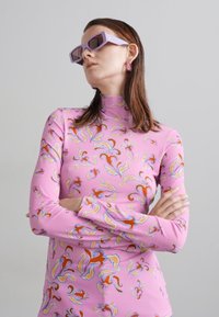 Haut à manches longues rose avec des motifs abstraits multicolores. Accompagné de lunettes de soleil carrées couleur lavande et de boucles d'oreilles pendantes.