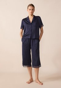 Jūras spēku satīna pidžamu komplekts ar īsām piedurknēm un pogu aizdari. Krūšturis un bikses ir dekorētas ar mežģīnēm, bet bikses ir apgrieztas līdz vidus teļiem.