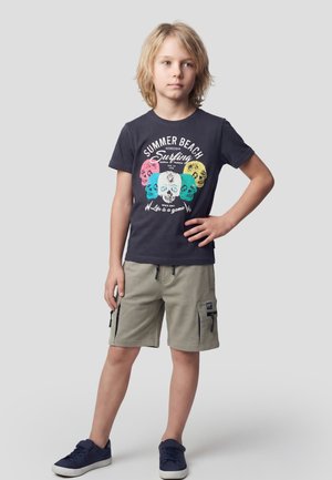 Niño rubio de pie con la mano en la cadera, vistiendo una camiseta azul marino con estampado de calaveras, pantalones cortos cargo beige y zapatillas azul marino sobre fondo gris.