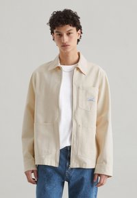 Beige canvasjas met een kraag, voorzak en knoopsluiting. Gemaakt over een wit shirt met blauwe spijkerbroek.