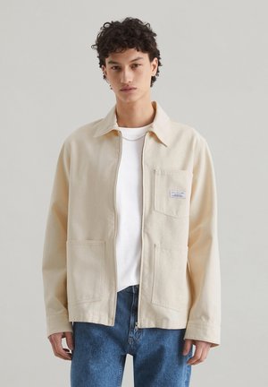 Beige canvas jacket with a collar, front pocket, and button closure. Worn over a white shirt with blue denim jeans.