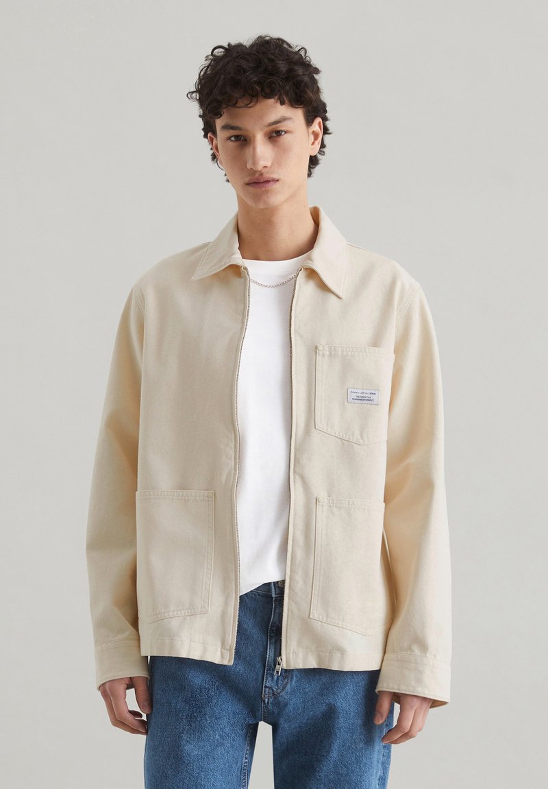 Beige canvasjas met een kraag, voorzak en knoopsluiting. Gemaakt over een wit shirt met blauwe spijkerbroek.
