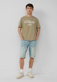Olijfgroen T-shirt met witte "s.Oliver" logo, lichtblauwe denim shorts en beige sneakers met witte accenten en lichtgekleurde sokken.