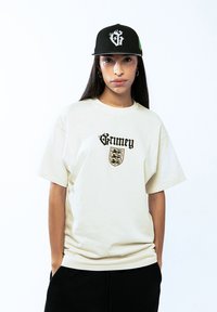 Camiseta oversized beige con el logo negro "Grimey" y diseño de escudo. Se lleva con una gorra negra que presenta una letra "G" estilizada y un acento verde.