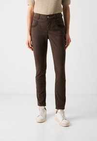Hnedé nohavice z bavlneného zmesi so slim fit strihom, prednými vreckami a prešívanými detailmi, skombinované s bielou obuvou na svetlom pozadí.