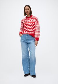 Suéter de punto rojo con patrón de corazones y acentos blancos, combinado con jeans de pierna ancha azul claro y zapatos oscuros. Textura suave, estilo casual.