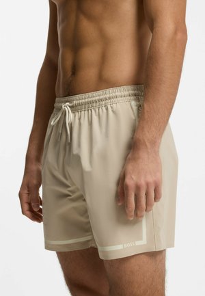 Mężczyzna noszący beżowe spodenki kąpielowe ze ściągaczem i logo BOSS na lewej dolnej nodze, widoczny tors i ramiona na białym tle.