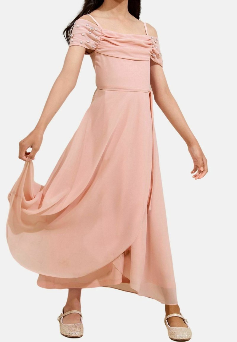 Robe en chiffon rose avec un corsage froncé, des manches courtes bouffantes ornées de perles et une jupe fluide. Portée avec des chaussures plates scintillantes.