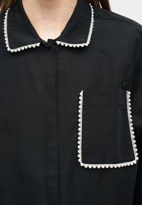 Camisa negra con cuello y bolsillo en pecho, ambos bordeados con un ribete de cuentas blancas redondas que parecen perlas.