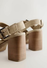 Sandales à talons carrés en daim beige avec brides réglables à boucle et talons empilés en bois sur un fond clair uni.