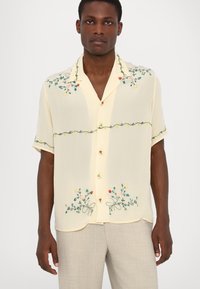 Camisa de manga corta de color amarillo claro con bordado floral en rojo, verde y amarillo. Presenta un cuello con muesca y cierre delantero con botones.