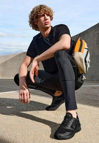 Schwarze Sportler-Sneaker aus strukturiertem Material mit Schnürsenkeln, getragen zu engen schwarzen Leggings und einem dunklen Poloshirt. Ein mehrfarbiger Rucksack ist sichtbar.