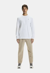 Biała koszula z długim rękawem z delikatnym logo, połączona z beżowymi joggerami o tapered kroju oraz szarymi butami sportowymi. Gładka tekstura i minimalistyczny design.