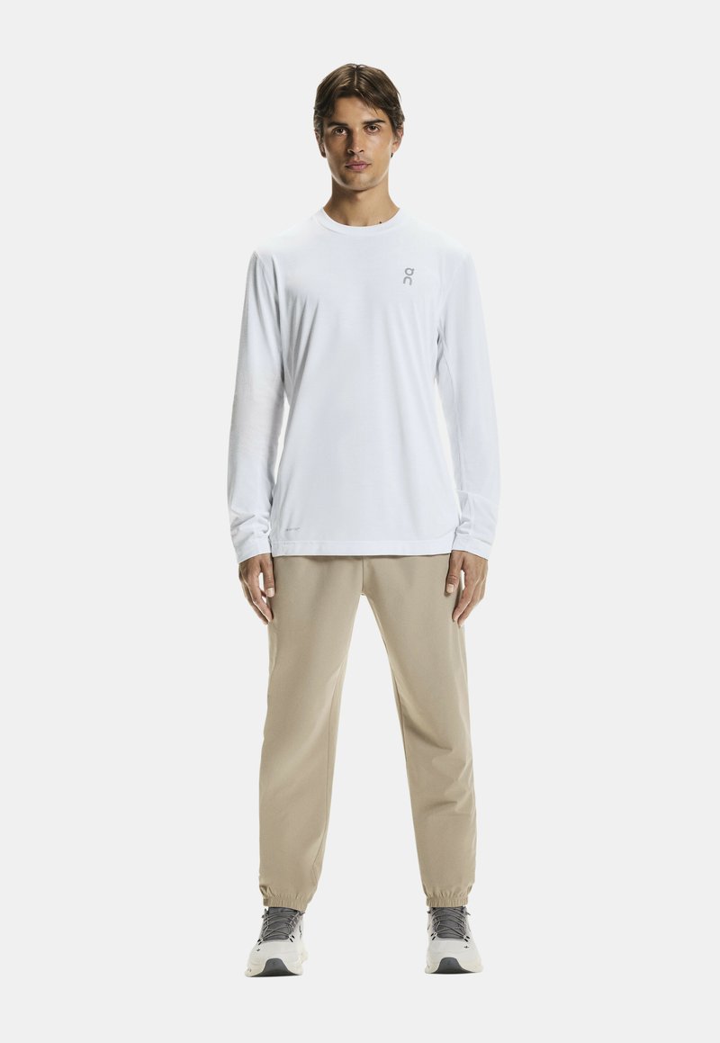 Biała koszula z długim rękawem z delikatnym logo, połączona z beżowymi joggerami o tapered kroju oraz szarymi butami sportowymi. Gładka tekstura i minimalistyczny design.