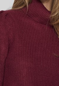 Gros plan sur une femme portant un pull en maille texturée couleur bordeaux avec un col montant, aux cheveux blonds longs partiellement visibles.