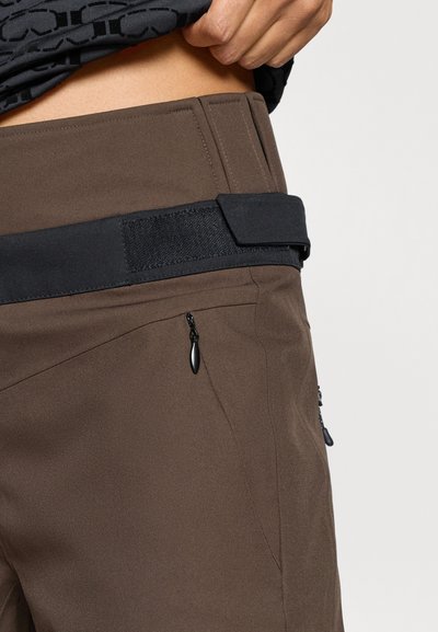 Pruunid püksid, millel on must reguleeritav vöökoht, tõmblukuga taskud ja sile tekstuur. Disain sisaldab nurgelisi õmblusi ja minimalistlikke detaile.