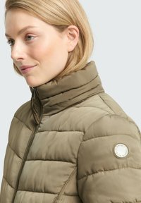 Kobieta z blond włosami nosząca jasnobrązową pikowaną kurtkę z wysokim kołnierzem i zamkiem, widoczna z profilu w lewo na jednolitym jasnym tle.