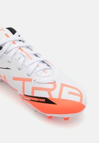 Baltas sintētisks futbola apavu pāris ar oranžiem akcentiem, ar strukturētu virsmu, melnu svītru un izteiktu logotipu. Šņores ir baltas.