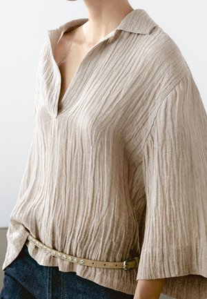Beige textured long-sleeve shirt with open collar tucked into dark pants, cinched with beige studded belt at waist.