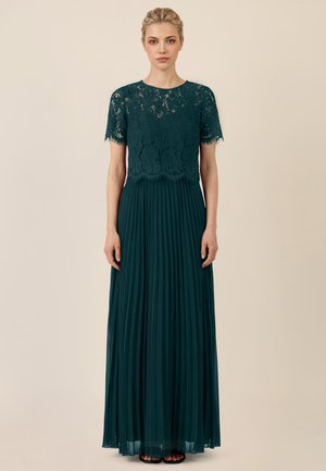 Frau trägt ein dunkelgrünes Kleid mit einem Spitzenoberteil mit kurzen Ärmeln und einem langen, plissierten Rock, steht vor einem schlichten beigen Hintergrund.
