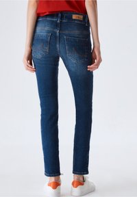 Jean en denim bleu skinny avec une taille mi-haute, comprenant deux poches arrière, des coutures orange et un patch de marque sur la ceinture.